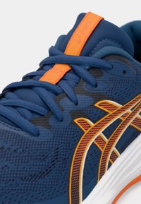 Μπλε αθλητικό παπούτσι τρεξίματος με υφασμάτινη επιφάνεια και υφή, πορτοκαλί και λευκές λεπτομέρειες, και επενδυμένο κολάρο. Διαθέτει κομψό σχεδιασμό με ορατά κορδόνια.