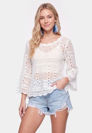 Ung kvinde med langt blondt hår iført en hvid hæklede top, revnede lyse denimshorts og blå fjerøreringe, smilende med hånden i lommen.