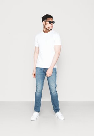 Witte T-shirt met korte mouwen gecombineerd met lichtblauwe jeans en witte sneakers. Het model draagt een zwart zonnebril en heeft kort, krullend haar.