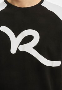 Juodas trumpomis rankovėmis marškinėliai, kuriuose centre yra didelis stilizuotas baltas logotipas ir kontrastingi balti akcentai ant pečių.