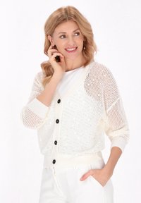 Weiße Strickjacke mit einem lockeren, durchbrochenen Design, schwarzen Knöpfen, gerippten Bündchen und einem verkürzten Saum. Getragen über einem weißen Oberteil.