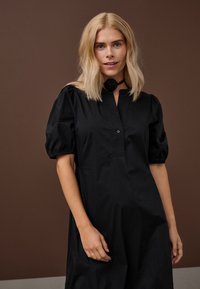 Šviesiaplaukė moteris juoda suknele su pūstomis rankovėmis ir juodu rožių chokeriu, stovinti prie rudų dažų fono.