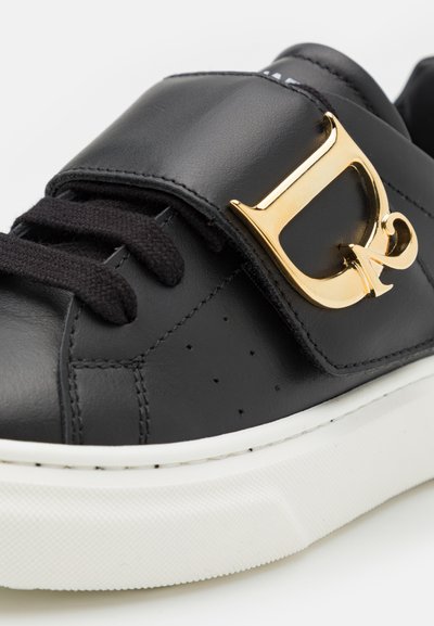 Sneaker in pelle nera con grande logo dorato, lacci neri, suola bianca texturizzata e perforazioni sulla punta per la traspirabilità.