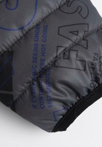 Sort quiltet stof med blå tekst og geometriske mønstre; har en struktureret overflade og en sort elastisk manchet ved bunden.