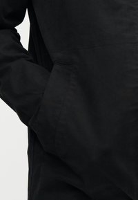 Το χέρι ενός ατόμου τοποθετημένο μέσα στην τσέπη ενός μαύρου μπουφάν κοτλέ, που δείχνει την λεπτομερή υφή και τις ραφές του υφάσματος.