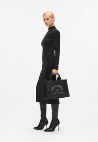 Sac en cuir noir avec logo argenté, design structuré et anses courtes, porté par un modèle dans une longue robe noire et des bottes stilettos jusqu'aux genoux.