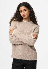 Ženska z dolgimi kodrastimi lasmi, oblečena v bež puhasto pulover z belim robom, ki se objema ob enobarvni beli ozadju.