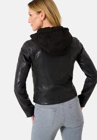 Giacca di pelle nera con cappuccio in camoscio. Design aderente, texture liscia e dettagli delle cuciture visibili. Abbinata a jeans in denim grigio chiaro.