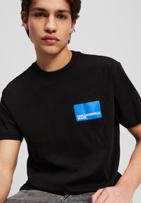 Melnā kokvilnas T-kreklā ir zils taisnstūra krūšu logs ar tekstu "KARL LAGERFELD JEANS"; īsas piedurknes, apaļa kakla izgriezums.