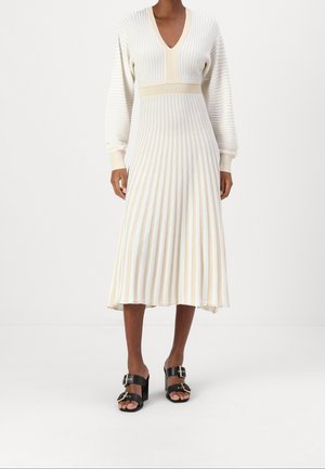 Žena nosící krémové plisované midi šaty s dlouhými rukávy a výstřihem do V, doplněné černými sandály na podpatku se zlacenými přezkami.