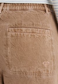Pantalones texturizados marrón claro con corte recto, que cuentan con un bolsillo trasero y un sutil logotipo de corazón bordado. Detalle de cintura elástica visible.