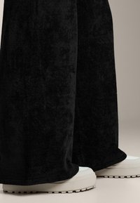 Čierne, plyšové široké nohavice s jemnou štruktúrou, v kombinácii s robustnými bielymi teniskami so štruktúrovanou podrážkou.