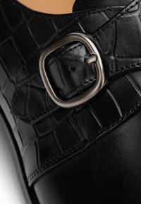 Schwarzer Lederschuh mit strukturiertem Krokodilmuster und einem metallischen Schnallenakzent auf dem Riemen. Glatte Oberfläche mit abgerundeter Form.