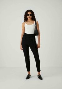 Calças pretas ajustadas com cintura alta e cinto, combinadas com uma blusa branca sem mangas e mules pretos. Os acessórios incluem uma pequena bolsa preta e óculos de sol.