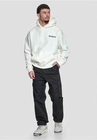 Hvit hettegenser med lomme foran og lilla logo, sammen med svarte cargobukser med sidelommer og grå sneakers.