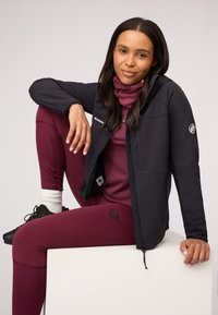 Black softshell jacket with hood, zippered pockets, and logo. Fitted maroon leggings and matching scarf. Textured fabric.