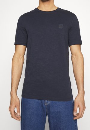 Homme portant un t-shirt bleu marine uni à manches courtes et un jean bleu sur un fond blanc uni.