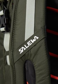 Grønn stoff skulderstropp med grå aksenter og hvit "SALEWA" logo. Strukturerte overflater med polstrede seksjoner for komfort og støtte.