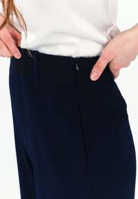 Marineblaue Hose mit glatter Textur, ausgestattet mit einem dezenten Reißverschluss am Bund und seitlichen Taschen für Funktionalität.