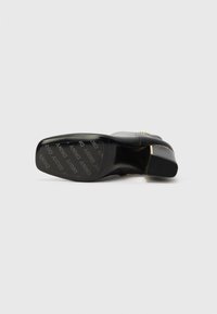 Black high-heeled shoe with a smooth leather upper, a pointed toe, and a gold accent on the heel. The sole features repeated branding.