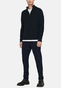 Pull en maille bleu marine avec un col zippé mi-hauteur, porté sur un t-shirt blanc. Assorti avec un pantalon foncé et des bottines en cuir noires.