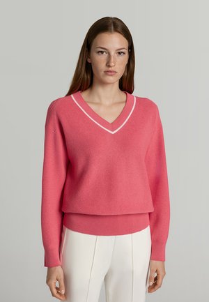 Ung kvinde med glat brunt hår iført en lyserød v-hals sweater med hvid kant og hvide bukser, stående mod en neutral baggrund.