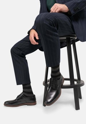 Svarta läderskor med runda tår och snörningsdesign, kombinerade med mörkrandiga strumpor. Modellen sitter på en svart pall, iklädd en randig kostym.