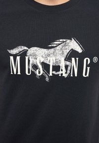 Černé tričko s opotřebovaným bílým grafikou skákajícího koně nad slovem "MUSTANG ®" v tučných, velkými písmeny.