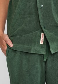 Groene badstof outfit met een knoopsluiting bovenstuk en broek met trekkoord. Opmerkelijke details zijn een zichtbaar label en een voorzak.