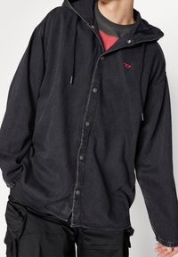 Man i en lös svart skjorta med knappar över en röd och svart t-shirt, ihop med svarta cargo-byxor, med handen i fickan.