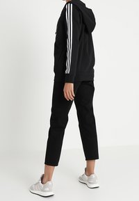 Svart hoodie med vit tre-stripesdesign på ärmarna, svarta byxor och ljusgrå sneakers med vita detaljer. Avslappnad sportklädsel.