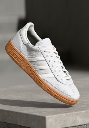 Witte suède Adidas sneaker met gumzool en drie witte strepen, schuin weergegeven op een betonnen oppervlak met een donkere, vage achtergrond.