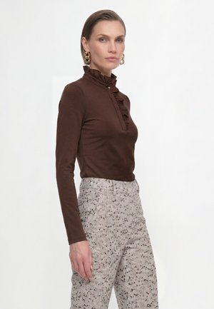 Woman wearing a brown long-sleeve top with ruffled collar and button detail paired with high-waisted light patterned pants and gold hoop earrings.