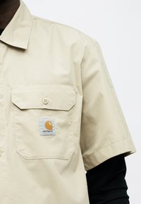 Μπεζ κοντομάνικο πουκάμισο με μπροστινή τσέπη με κουμπιά και λογότυπο Carhartt, φορεμένο πάνω από μαύρο μακρυμάνικο τοπ.