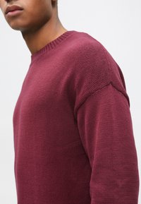 Bordowy sweter z dzianiny z okrągłym dekoltem, z prążkowanymi mankietami i fakturowaną tkaniną. Zbliżenie na detal na ramieniu.