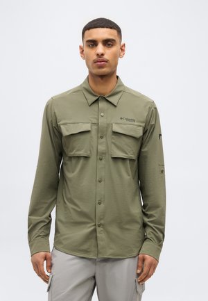 Egy férfi olívazöld, gombos ingben, mellzsebbel és világosszürke terep nadrágban, egyszerű világos háttér előtt áll.