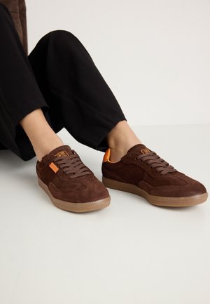 Brown suede sneakers with a rounded toe, textured upper, and orange accents. Features flat laces and a rubber sole.