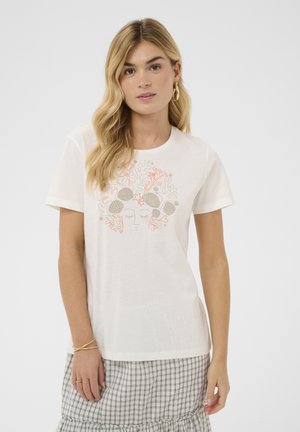 Ung kvinde med langt blondt hår iført hvid T-shirt med koral- og muslingemotiv og ternet nederdel, stående foran en ensfarvet baggrund.