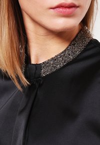 Detailní záběr na krk a rty ženy v černém saténovém oděvu s dekorativním korálkovým límcem.