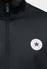 Μαύρο μπουφάν με φερμουάρ από υφαντό ύφασμα. Διαθέτει στρογγυλό, λευκό λογότυπο με μπλε αστέρι και κόκκινο περίγραμμα που γράφει "Converse Chuck Taylor".