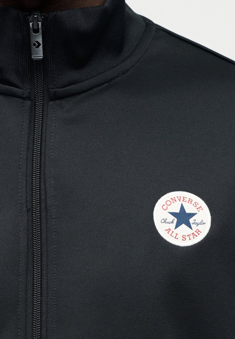 Schwarze Reißverschlussjacke aus strukturiertem Stoff. Mit rundem, weißem Logo mit einem blauen Stern und rotem Rand, auf dem "Converse Chuck Taylor" steht.