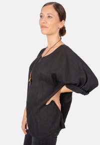 Schwarzes Leinenoberteil mit lockerer Passform, weiten Ärmeln und rundem Ausschnitt, das eine lange Kette mit bunten Akzenten hat.