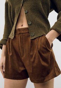 Shorts marroni in suede a vita alta indossati con un cardigan verde oliva a coste con bottoni, una mano in tasca, tagliati a metà coscia e vita.