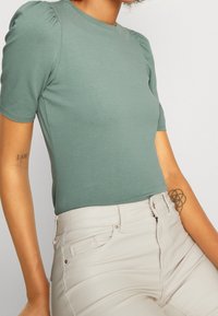 Grön figurnära topp med korta puffärmar, parat med krämfärgade byxor med blixtlåsförslutning och subtila sömdetaljer.