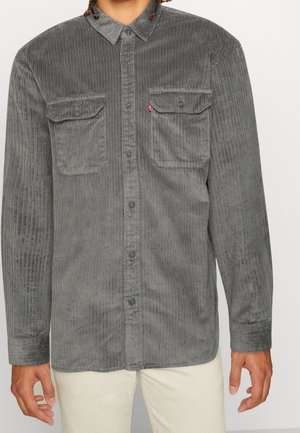 Mężczyzna ubrany w szarą sztruksową koszulę z dwiema zapinanymi na guziki kieszeniami na piersi oraz kremowe spodnie.