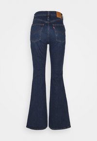 Jean en denim bleu foncé taille haute avec jambes évasées, deux poches arrière, un patch en cuir marron sur la ceinture, et une petite étiquette rouge sur la poche droite.