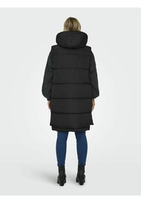 Gilet rembourré noir avec un col haut et une capuche, présentant un large design matelassé et une forme allongée, porté par-dessus un pull texturé sombre.