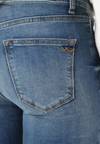 Kék farmer, textúrált denim felülettel, hátul kerek kialakítású zsebbel és kontrasztos sárga varrással.
