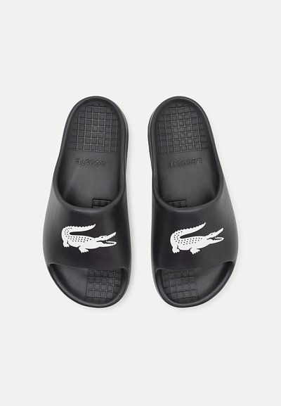 Sandales en slide noires avec une semelle texturée, un design à bout ouvert et un logo crocodile blanc sur le dessus. Fabriquées en matériau synthétique durable.