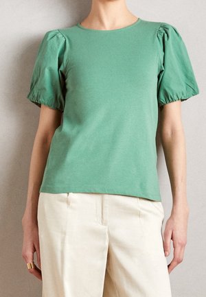 Persoon draagt een groene top met korte mouwen en opgepofte schouders en een beige broek met hoge taille tegen een effen achtergrond.